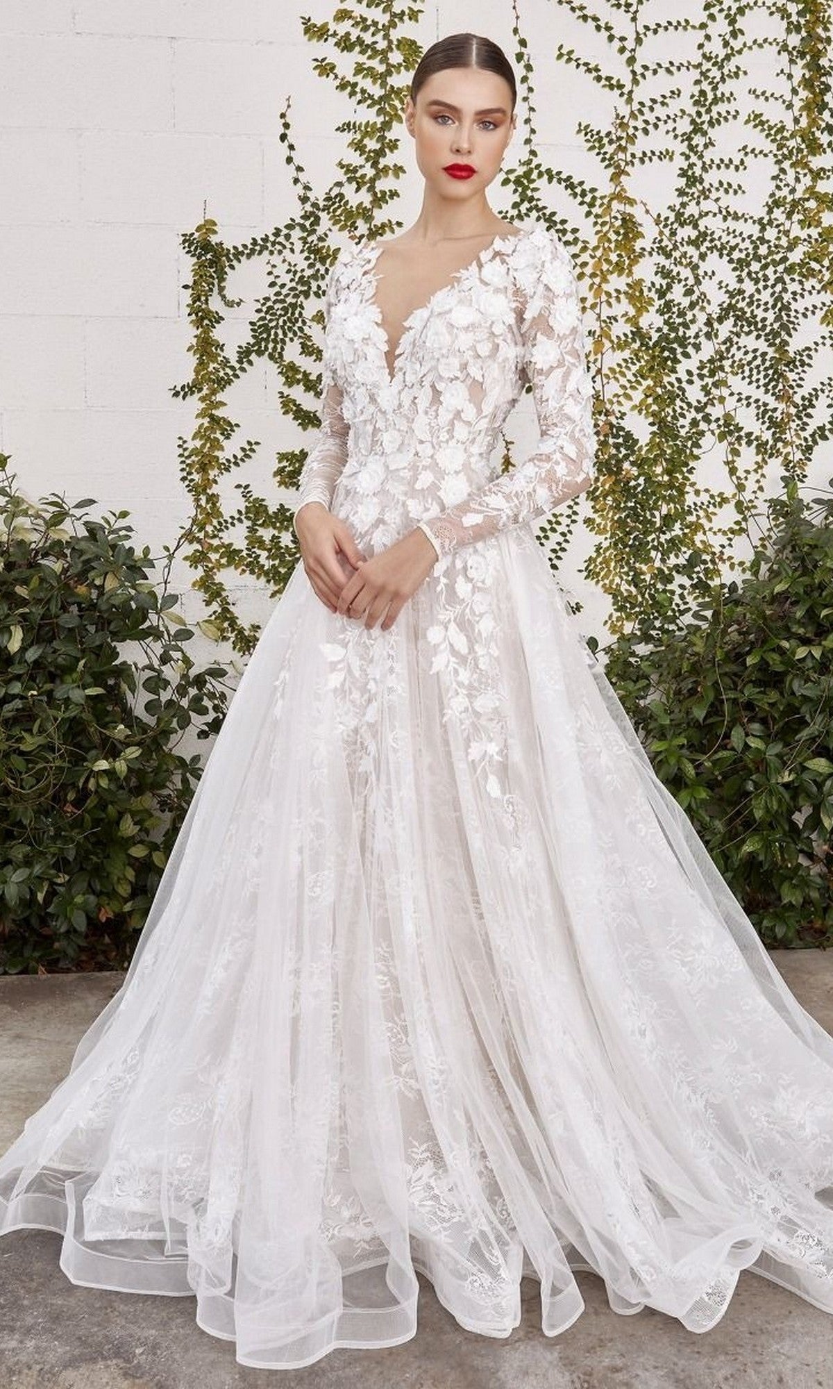 A woman wearing the Andrea and Leo A1067WC long formal dress, featuring elegant long sleeves and white floral lace appliques, stands outdoors in front of a vine-covered wall.